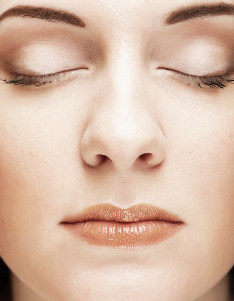 Un primer plano facial de una mujer con piel lisa, cejas oscuras, ojos cerrados y lápiz labial oscuro de color nude.